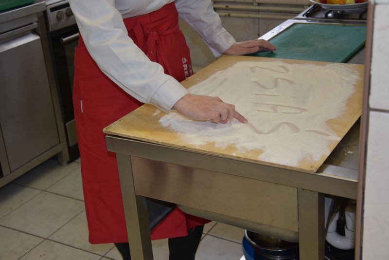 Uczeń technikum gastronomicznego wykonuje palcem na wysypanej mąką stolnicy skróconą nazwę szkoły czyli ZSiAŚ