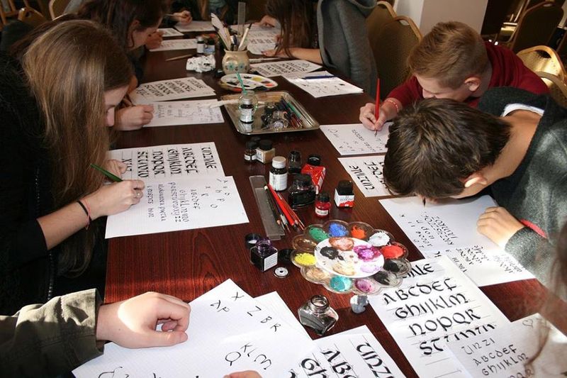 Grupa uczniów przy stole konferencyjnym ćwiczy kaligrafię liter za pomocą pióra oraz atramentu. Pośrodku stołu znajdują się potrzebne przybory – pióra, pędzle, małe butelki z atramentem oraz paleta kolorów